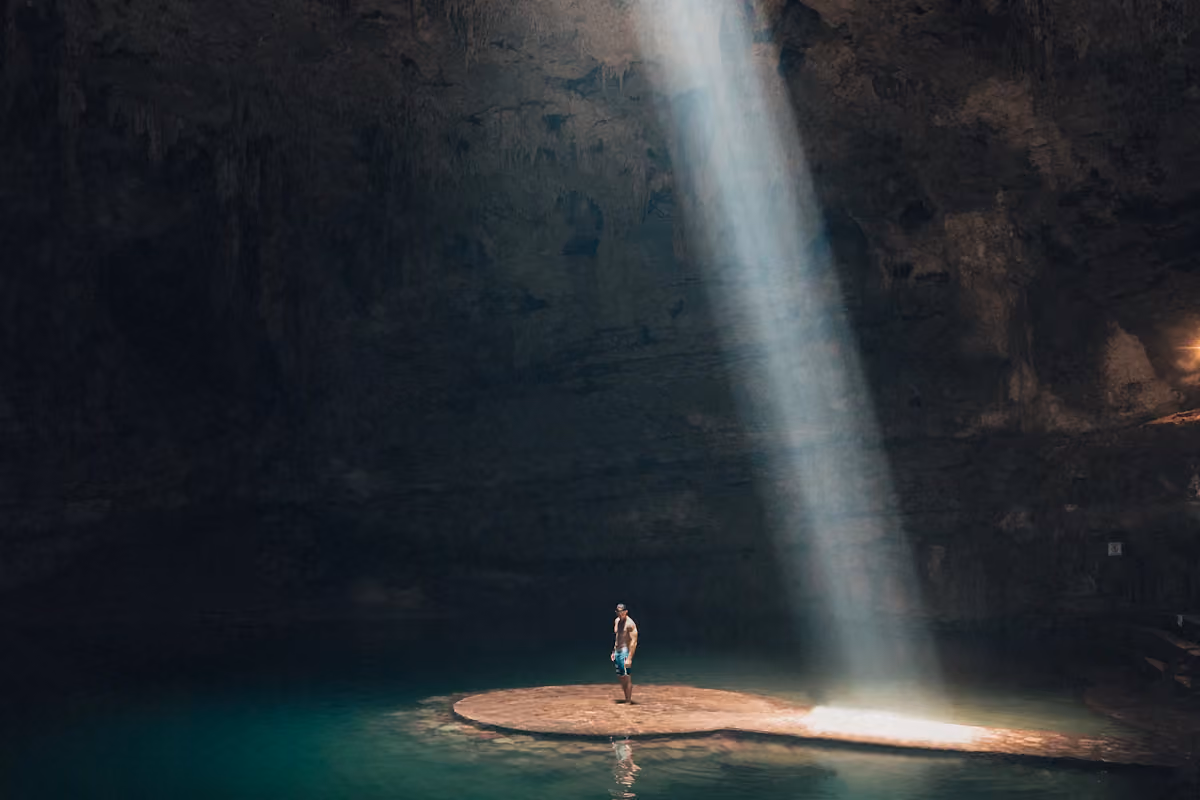 Sunlight entering a limestone cavern above a person beside blue water